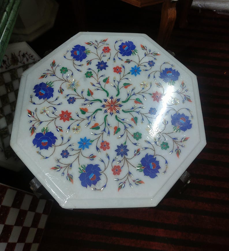 May include: Octagonal white marble table top with intricate floral design inlaid with colorful stones. The table top has a polished finish and is surrounded by a wooden frame.