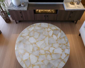 Puede incluir: Una mesa de centro redonda con una superficie de cuarzo blanco y dorado. La mesa es el punto focal de una sala de estar moderna, con un mueble de entretenimiento de madera y un jarrón blanco en el fondo.