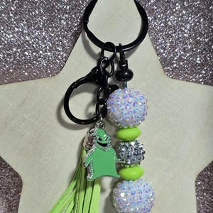 May include: A black keychain with a green tassel and a green Oogie Boogie charm. The keychain features iridescent and silver beads with lime green accents. The keychain is attached to a black clasp and ring.