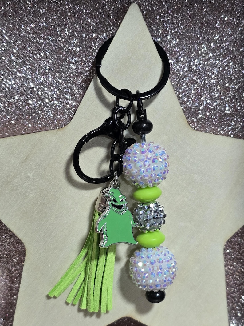May include: A black keychain with a green tassel and a green Oogie Boogie charm. The keychain features iridescent and silver beads with lime green accents. The keychain is attached to a black clasp and ring.