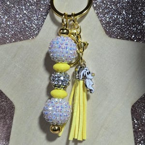 May include: A gold-toned keychain with a beaded design. The keychain features iridescent white and silver beads, yellow accents, and a yellow tassel. A small charm of a cartoon ghost is attached.
