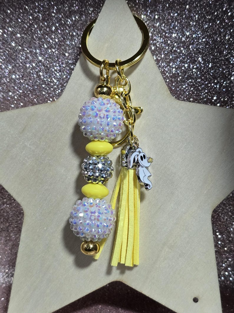 May include: A gold-toned keychain with a beaded design. The keychain features iridescent white and silver beads, yellow accents, and a yellow tassel. A small charm of a cartoon ghost is attached.