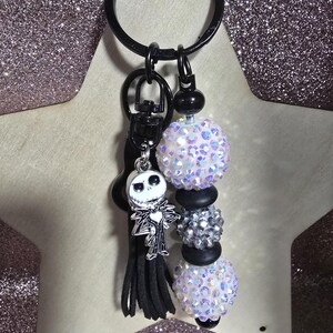 May include: A black keychain with a Jack Skellington charm, black tassel, and beaded accents. The keychain features iridescent white and silver beads, and black beads. The background is a glittery pink.