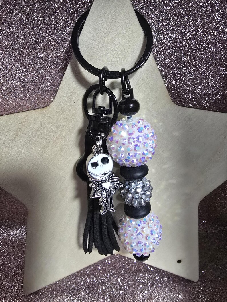 May include: A black keychain with a Jack Skellington charm, black tassel, and beaded accents. The keychain features iridescent white and silver beads, and black beads. The background is a glittery pink.