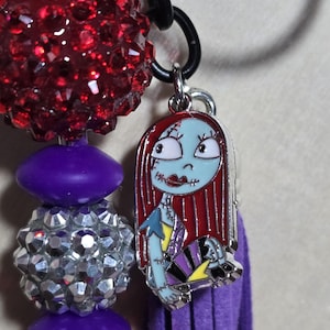 May include: A beaded keychain featuring a charm of Sally from The Nightmare Before Christmas. The charm is attached to a purple tassel and a chain with red and silver beads.