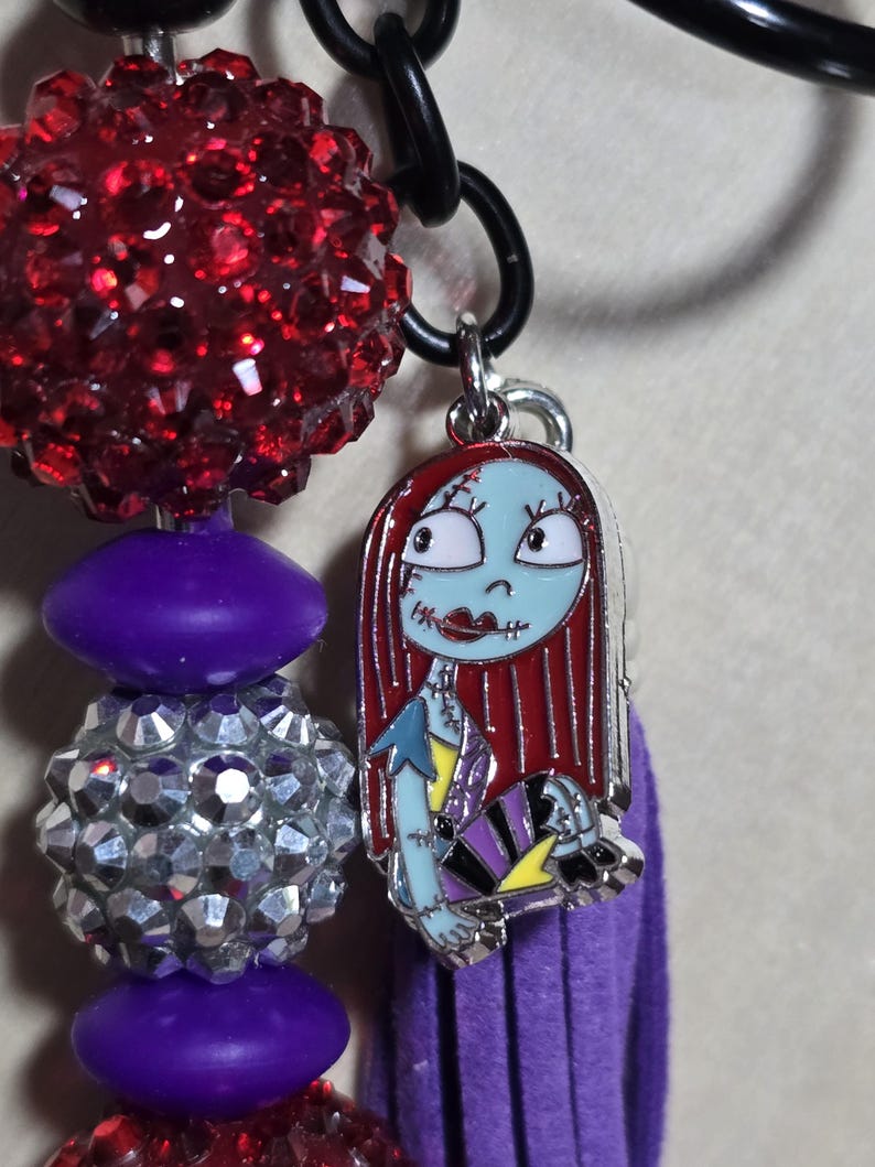 May include: A beaded keychain featuring a charm of Sally from The Nightmare Before Christmas. The charm is attached to a purple tassel and a chain with red and silver beads.