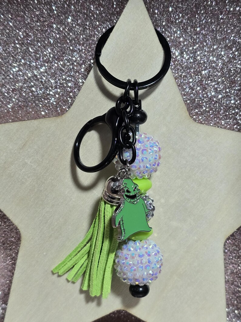 May include: A black keychain with a green tassel and a green Oogie Boogie charm. The keychain features two white, sparkly beaded balls and a black clasp. The charm is a green character from the movie, The Nightmare Before Christmas.