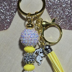 May include: A gold keychain with a beaded design. The keychain features iridescent white and silver beads, yellow beads, and a yellow tassel. Charms include a silver crown and a white ghost-like figure.