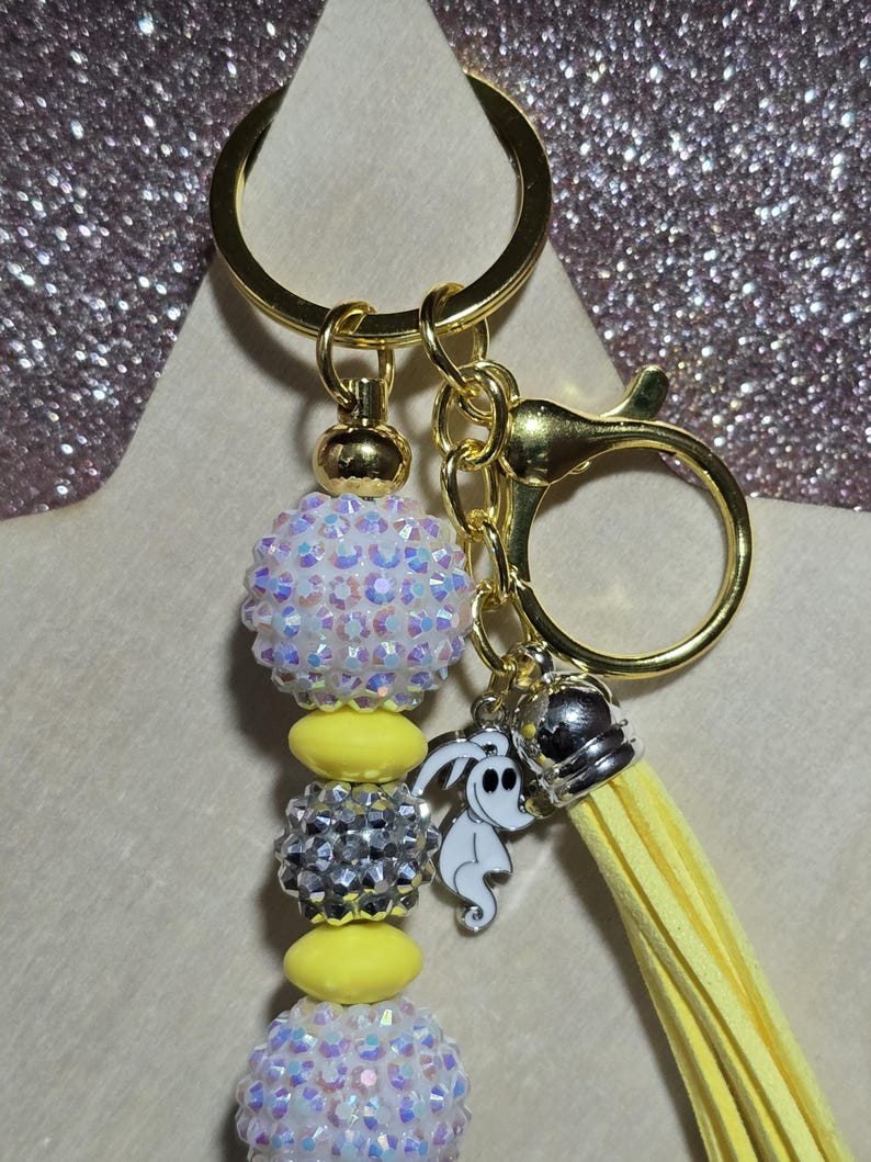 May include: A gold keychain with a beaded design. The keychain features iridescent white and silver beads, yellow beads, and a yellow tassel. Charms include a silver crown and a white ghost-like figure.