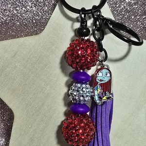 May include: A black keychain with a beaded design and a charm. The keychain features red rhinestone beads, purple beads, and a silver rhinestone bead. A purple tassel and a Sally charm are also attached.