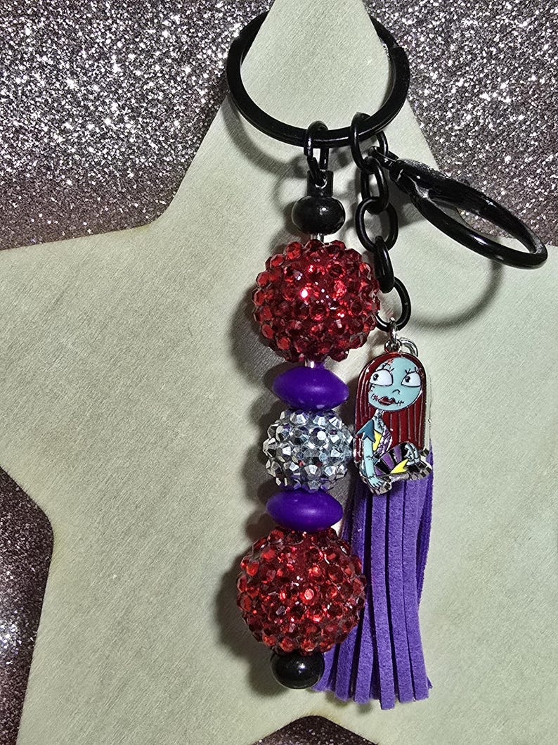 May include: A black keychain with a beaded design and a charm. The keychain features red rhinestone beads, purple beads, and a silver rhinestone bead. A purple tassel and a Sally charm are also attached.