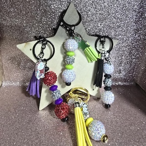 May include: A collection of keychains with decorative beaded accents and tassel details. The keychains feature various colors, including red, purple, silver, and yellow. Some keychains have character charms.
