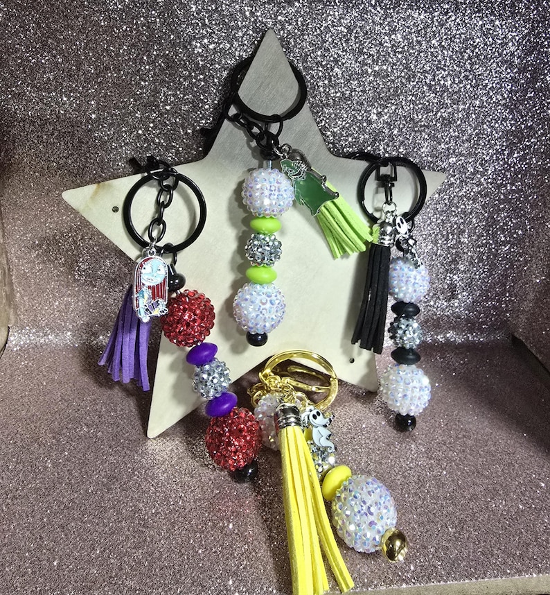 May include: A collection of keychains with decorative beaded accents and tassel details. The keychains feature various colors, including red, purple, silver, and yellow. Some keychains have character charms.