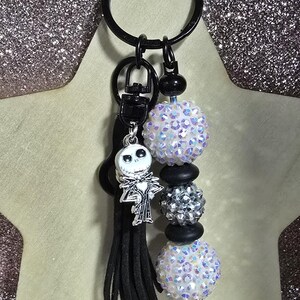 May include: A black keychain with a Jack Skellington charm, black tassel, and beaded accents. The beaded accents include iridescent and silver beads, and black spacers. The keychain is attached to a black key ring.