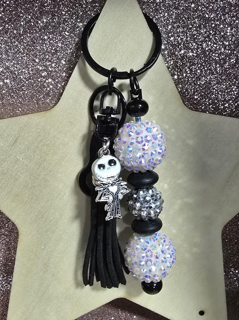 May include: A black keychain with a Jack Skellington charm, black tassel, and beaded accents. The beaded accents include iridescent and silver beads, and black spacers. The keychain is attached to a black key ring.