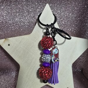 May include: A keychain featuring a charm of Sally from The Nightmare Before Christmas, with a purple tassel and beaded accents. The keychain includes red and silver glittery beads, and a black key ring.