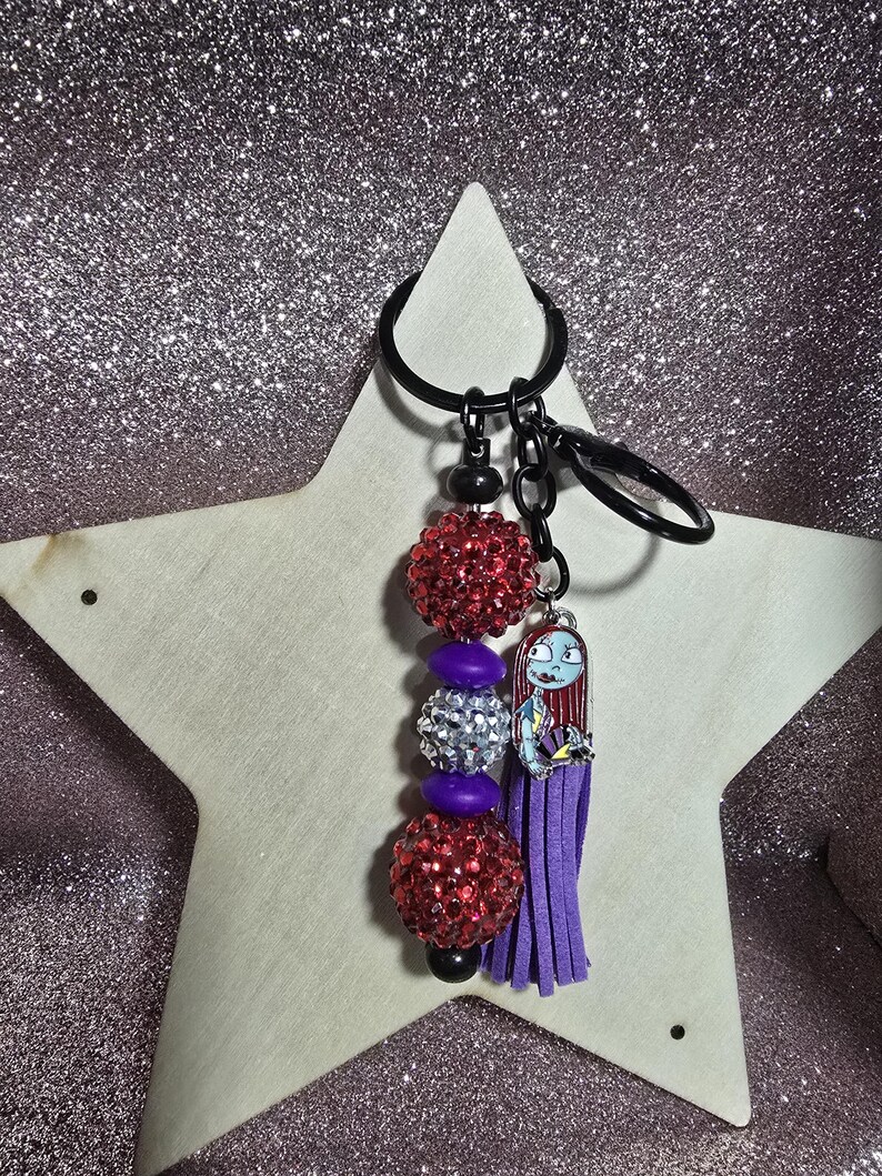 May include: A keychain featuring a charm of Sally from The Nightmare Before Christmas, with a purple tassel and beaded accents. The keychain includes red and silver glittery beads, and a black key ring.