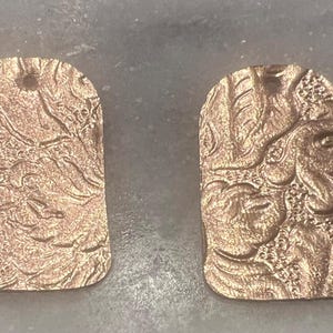 Könnte beinhalten: Zwei rechteckige Anhänger mit abgerundeten Oberkanten, jeweils mit einer metallischen Kupferoberfläche und einem erhabenen, komplizierten Blumenmuster. Jeder Anhänger hat ein kleines kreisförmiges Loch oben zur Befestigung.