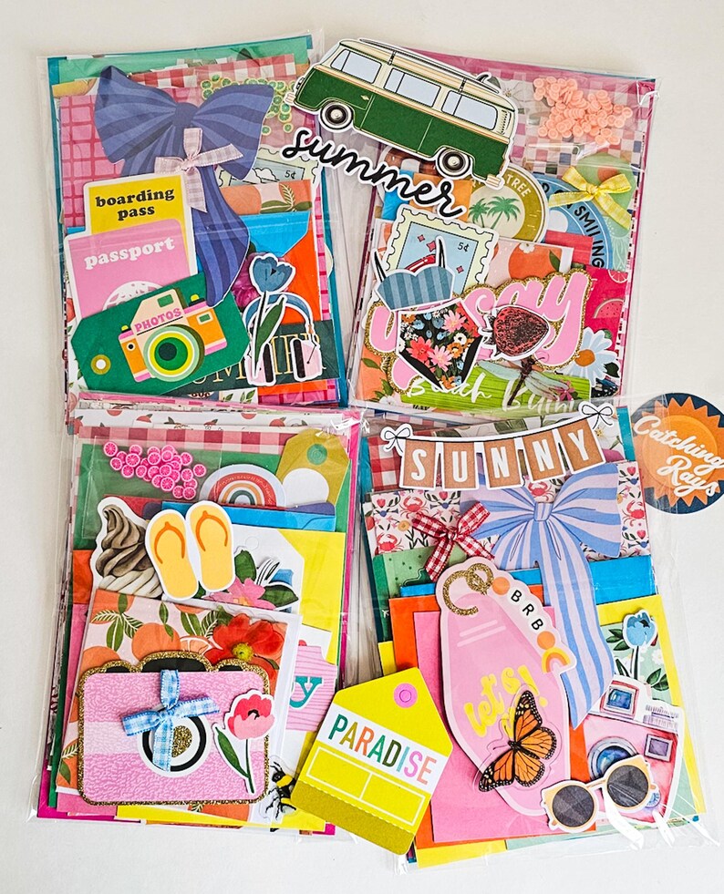 May include: Four colorful scrapbook kits with summer-themed embellishments. Each kit includes decorative paper, stickers, and die-cuts featuring travel motifs like cameras, passports, and a green van. The word "summer" is visible on one kit.