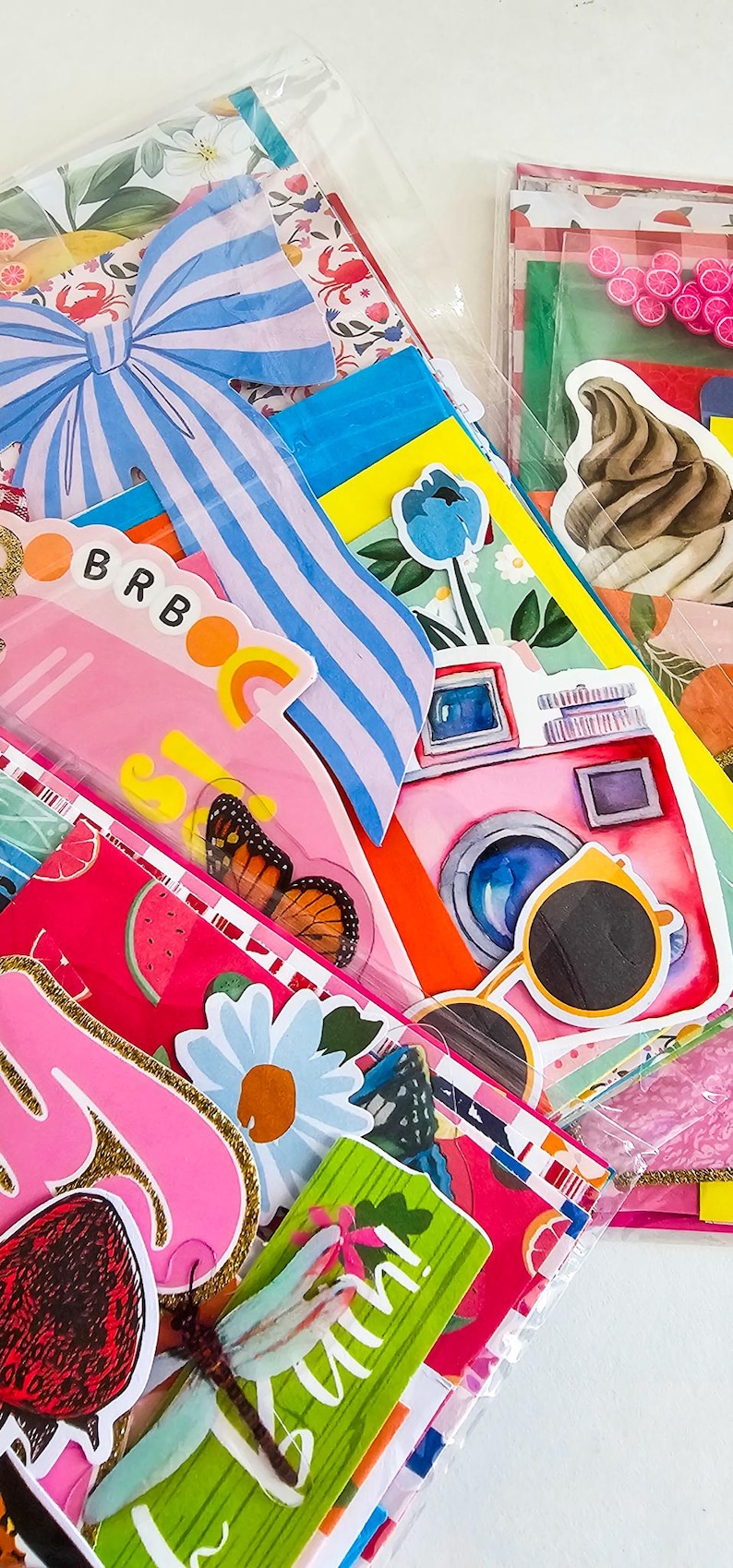 May include: A collection of colorful stickers and decorative paper items. The image features a pink camera, sunglasses, a butterfly, and the text "BRB". Other designs include a blue bow, flowers, and fruit.