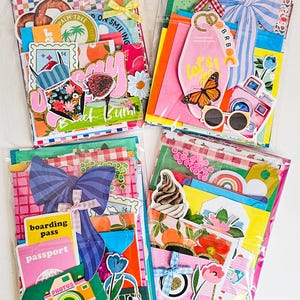May include: Four colorful scrapbook kits in clear plastic packaging. Each kit features decorative paper, stickers, and embellishments with travel and summer themes. Designs include bows, butterflies, cameras, and the text "Beach Bum."