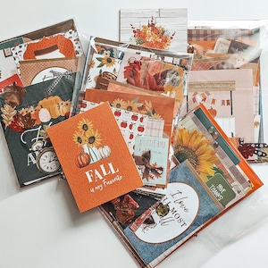 May include: An assortment of fall-themed paper crafts, including cards, patterned papers, and embellishments. The color palette features oranges, greens, and browns, with images of pumpkins, sunflowers, and the text "FALL is my Favorite".