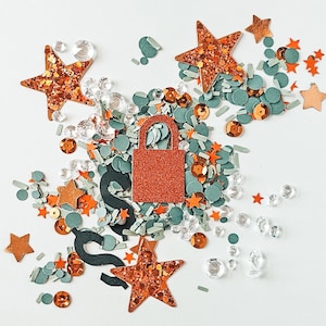 May include: An assortment of decorative elements including orange glitter stars, teal circles, clear gems, and orange sequins. A glittery orange padlock shape is centered, with dark green swirls. The arrangement is on a white background.