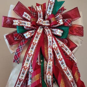 May include: A festive Christmas tree bow featuring layers of red, green, and gold ribbons. The bow includes ribbons with a nutcracker soldier pattern, plaid, and glitter accents. A large, cream-colored bow forms the base.