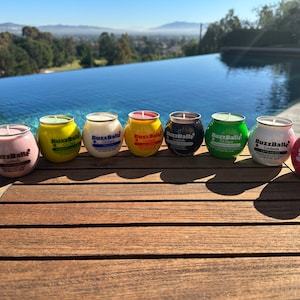 May include: A collection of seven colorful BuzzBallz Cocktails and Chillers, each in a round, metal-rimmed container. The containers are various colors, including pink, yellow, white, red, black, and green, with labels and text. The background features a pool and a scenic view.