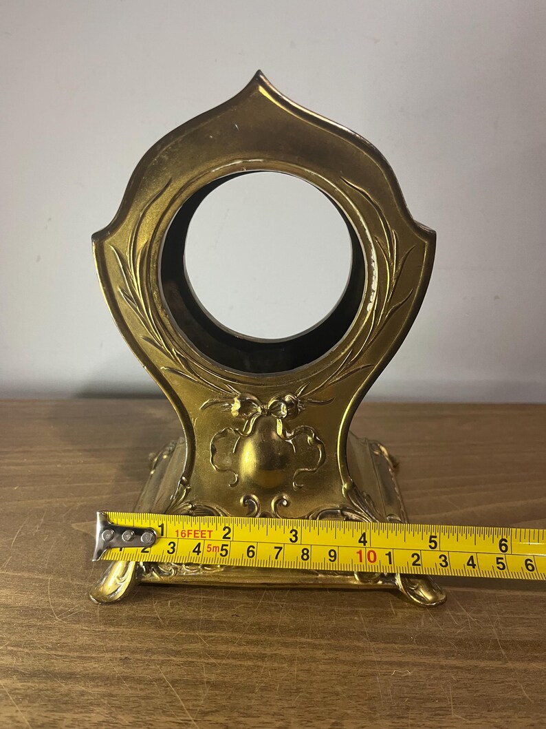 May include: A gold-coloured metal stand with an ornate design and a round opening in the centre. The stand is approximately 10 inches wide.