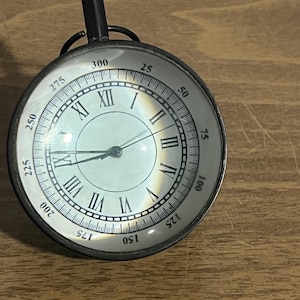 May include: A black metal clock with Roman numeral markings and a white face. The clock has a magnifying glass over the face.