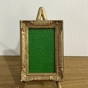 May include: A small, gold-colored easel-style picture frame with an ornate, detailed border. The frame has a green backing and is supported by three legs. The top of the frame has a decorative arch.