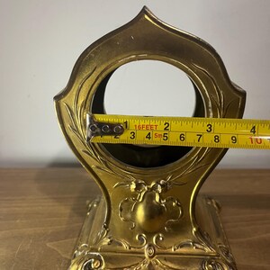 May include: A gold-coloured metal clock stand with an ornate design. The stand has a round opening in the top and a square base. The stand is 11.4 cm tall.