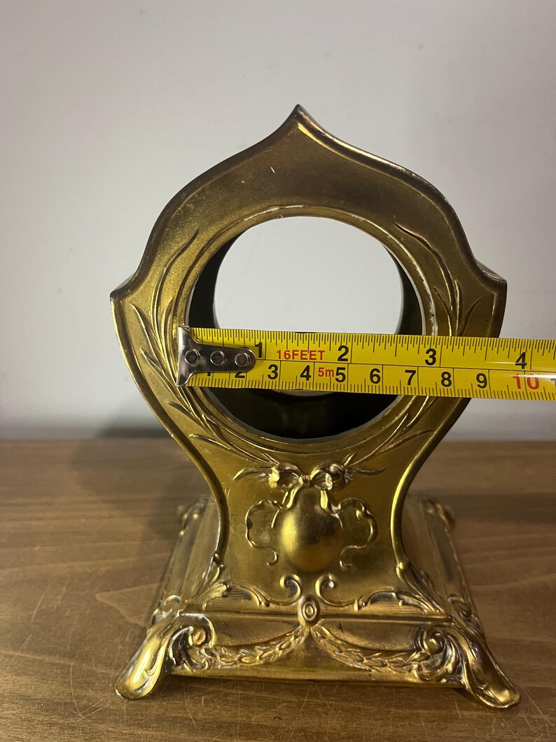 May include: A gold-coloured metal clock stand with an ornate design. The stand has a round opening in the top and a square base. The stand is 11.4 cm tall.