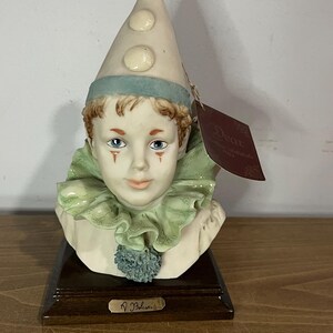 May include: A porcelain figurine of a clown with a blue and white hat, green ruffled collar, and blue eyes. The figurine is on a wooden base with the artist's signature "J. Beltran".