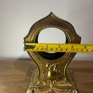 May include: A gold-coloured metal stand with an ornate design. The stand has a round opening at the top and a base with three legs. The stand is approximately 25 cm tall.