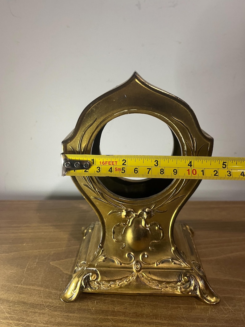 May include: A gold-coloured metal stand with an ornate design. The stand has a round opening at the top and a base with three legs. The stand is approximately 25 cm tall.