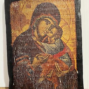 May include: A rectangular wooden icon featuring a painted depiction of a figure holding a child against a gold background. The artwork has a distressed, cracked texture and is framed by a thick, dark, textured border with visible wear.