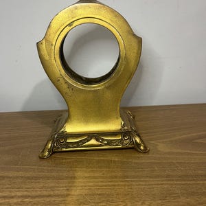 May include: Gold-toned metal clock stand with an ornate base and a circular opening for a clock face.