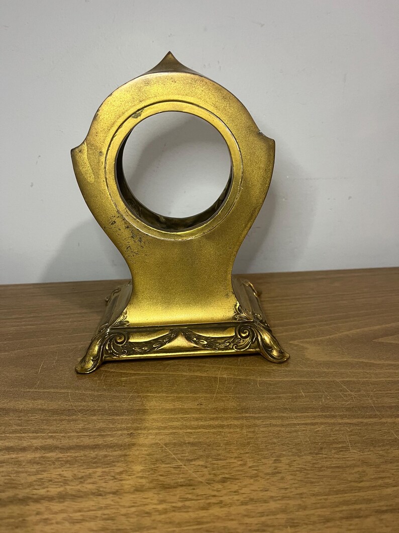 May include: Gold-toned metal clock stand with an ornate base and a circular opening for a clock face.