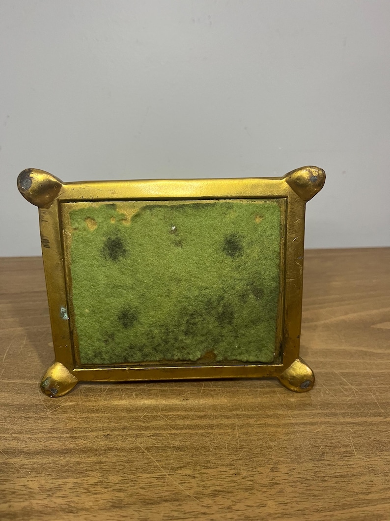 May include: A gold-coloured metal stand with a green felt bottom. The stand has a square base with rounded corners and four feet.