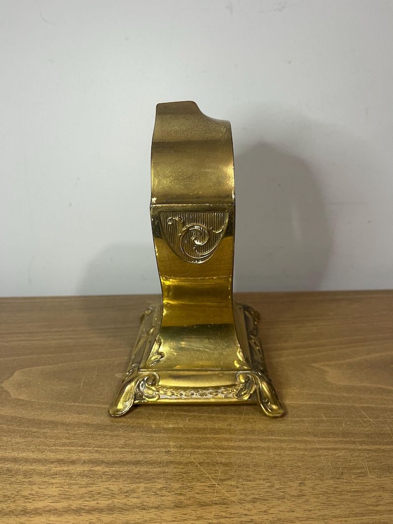 May include: A gold-toned metal decorative stand with an ornate design. The stand has a curved top and a square base with intricate details.