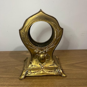 May include: A gold-coloured metal clock stand with an ornate design. The stand has a round opening in the centre and a square base with intricate details.