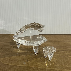 May include: A clear, crystal-like decorative piano with three pointed legs and a matching stool. The piano has an open lid and is set on a wooden surface. The piano and stool are made of faceted glass.