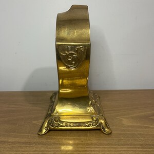 May include: A gold-toned metal clock with an ornate design. The clock has a square base with intricate details and a curved top.