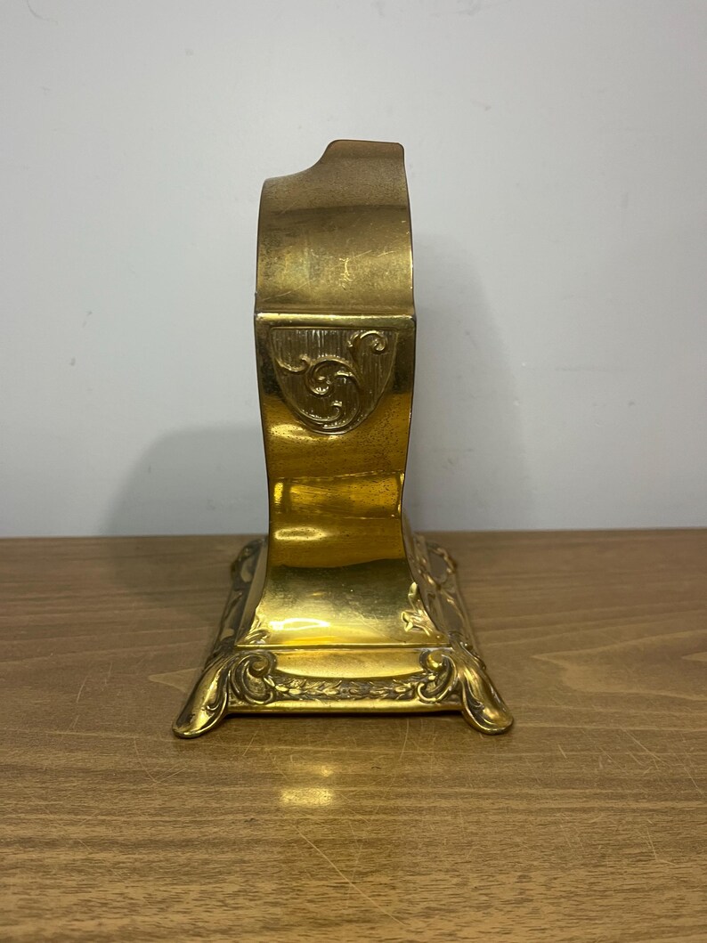 May include: A gold-toned metal clock with an ornate design. The clock has a square base with intricate details and a curved top.