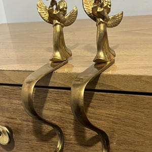 May include: Two brass-colored Christmas stocking holders. Each holder features a detailed angel playing a trumpet atop a curved, decorative hook. The angels have detailed wings and flowing robes. The hooks are designed to hang over a mantel or shelf.
