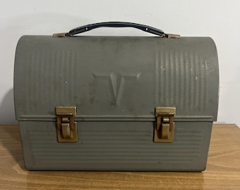 May include: A vintage green metal lunchbox with a black handle and two brass latches. The lunchbox has a raised letter "V" on the front.