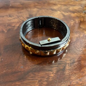 May include: A hinged bangle bracelet featuring a dark gunmetal finish on the interior and a row of gold-toned pyramid studs along the exterior. The bracelet includes a rectangular clasp mechanism with a small gold-colored spherical accent.