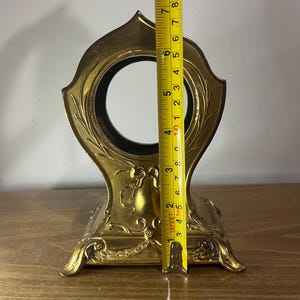 May include: A gold-coloured metal clock stand with an ornate design. The stand is approximately 15 cm tall.
